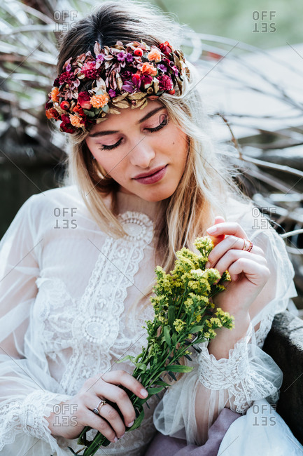 Beautiful young woman in white dress and floral wreath touching bouquet of small yellow flowers while resting in garden on wedding day