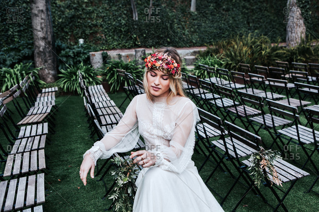 High angle of beautiful young female in elegant white dress and flower wreath closing eyes and sitting on empty chairs before wedding ceremony in garden