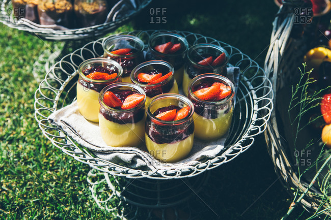 Composition of fresh tasty desserts decorated with strawberry served in glass jars placed on round metal tray green lawn in garden