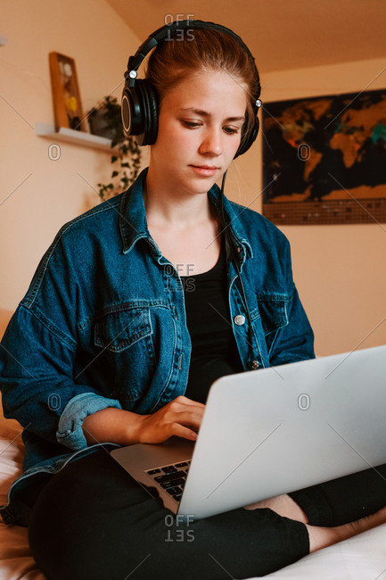 Joyful pensive female in headphones and casual clothes focusing on screen with interest and using netbook while sitting with legs crossed on soft bed in cozy modern apartment