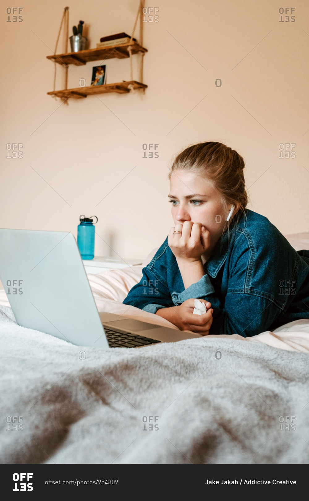 Thoughtful displeased female crying and using facial paper while lying down on bed watching sad movie on laptop against beige wall in light apartment