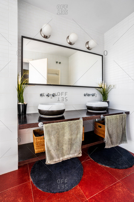 Modern bathroom with lamps and large mirror placed over stylish counter with sinks and cozy round carpets on red floor