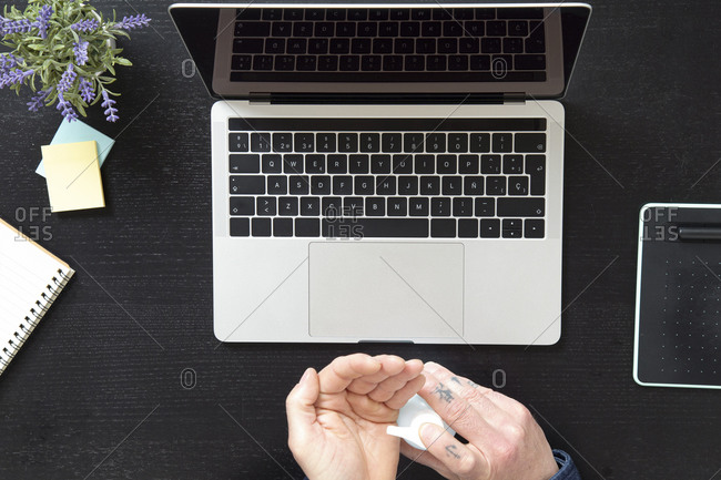 From above crop male treating hands with sanitizer for protection from coronavirus while working on laptop at home during quarantine