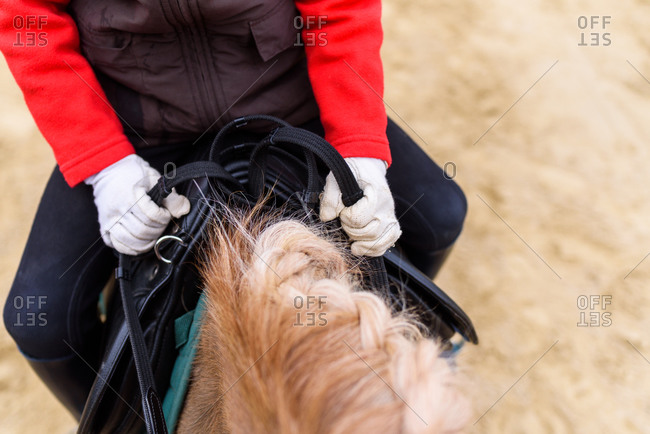 From above anonymous little jockey grasping reins and riding pony with braided man on sandy ground of paddock
