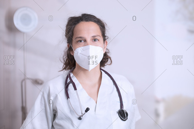 Positive young female doctor in white uniform and stethoscope wearing protective medical mask standing in modern medical office and looking at camera