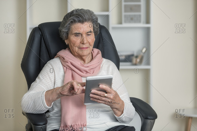 Positive senior woman in pink scarf smiling while sitting on comfortable chair in cozy room at home