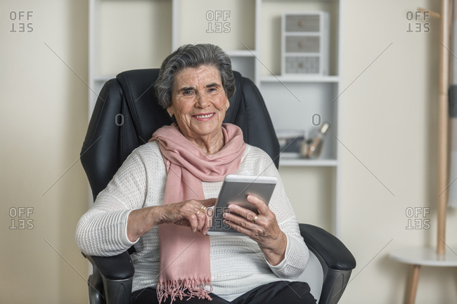 Positive senior woman in pink scarf smiling and looking at camera while sitting on comfortable chair in cozy room at home