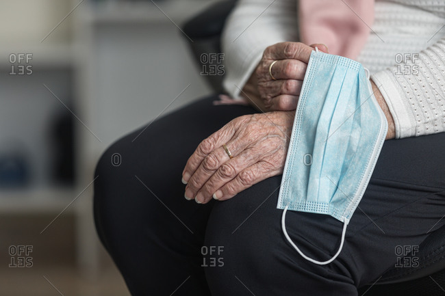 Anonymous elderly female holding medical mask while sitting on chair at home during coronavirus pandemic