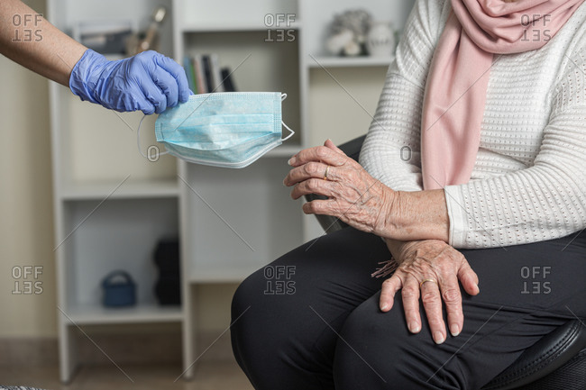 Crop unrecognizable social worker in latex gloves giving a medical mask to a elderly female on chair while taking care of pensioner during coronavirus pandemic at home
