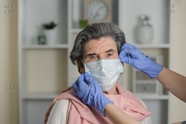 Crop anonymous social worker in latex gloves putting on medical mask on elderly female on chair while taking care of pensioner during coronavirus pandemic at home