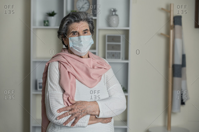 Elderly woman in medical mask and pink scarf looking at camera with arms crossed while staying at home during coronavirus pandemic