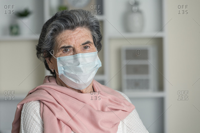 Elderly woman in medical mask and pink scarf looking at camera while staying at home during coronavirus pandemic