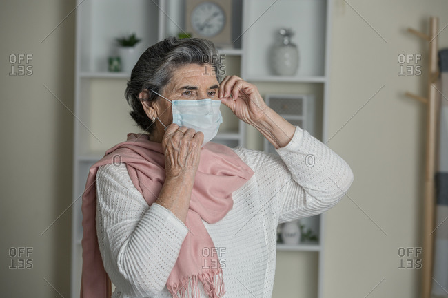 Elderly woman in medical mask and pink scarf looking away while staying at home during coronavirus pandemic