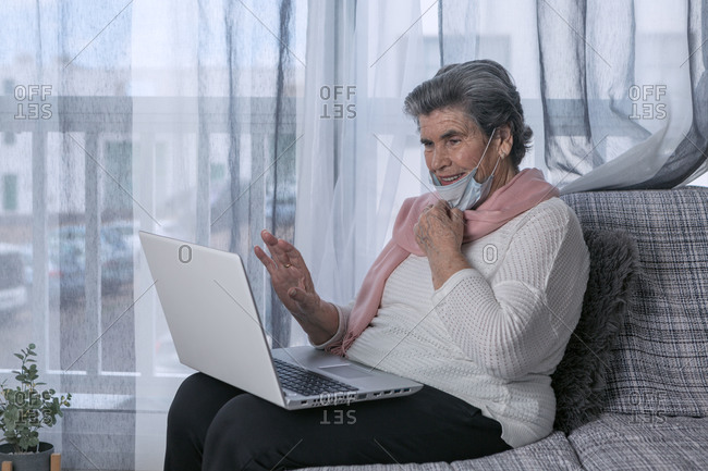 Senior female in medical mask sitting on couch and doing video call through laptop while staying at home during coronavirus pandemic