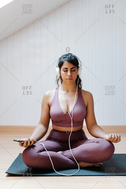 Serene fit female in sportswear listening to relaxing melody with headphones and smartphone while practicing meditation during yoga training at home