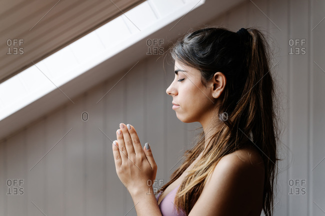 Tranquil fit female in sportswear with clasped hands practicing yoga meditation in modern light room in morning