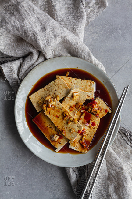 Top view of tofu slices with fragrant pieces of garlic marinating in soy sauce in white deep plate while standing on kitchen towel on gray table with steel  chopsticks