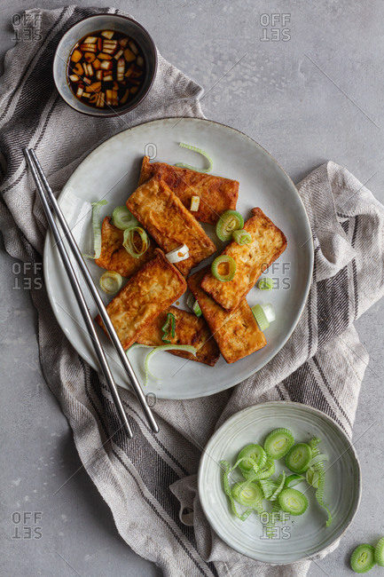 Top view of delicious vegetarian meal fried tofu marinating in soy sauce with garlic and serving in white plate sprinkling with pieces of green onion standing on kitchen towel on gray table with steel Chinese chopsticks
