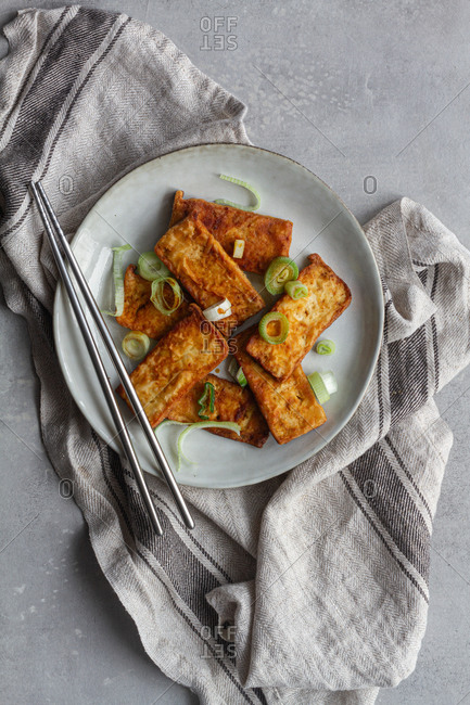 Top view of tofu slices with fragrant pieces of garlic marinating in soy sauce in white deep plate while standing on kitchen towel on gray table with steel chopsticks