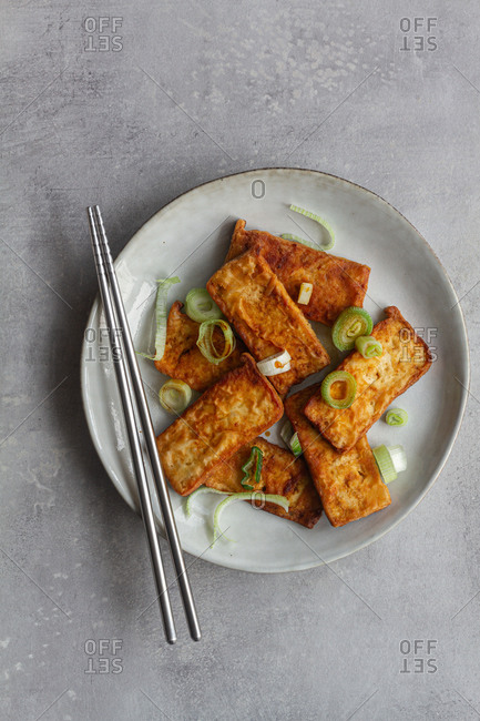 Top view of tofu slices with fragrant pieces of garlic marinating in soy sauce in white deep plate while standing on kitchen towel on gray table with steel chopsticks