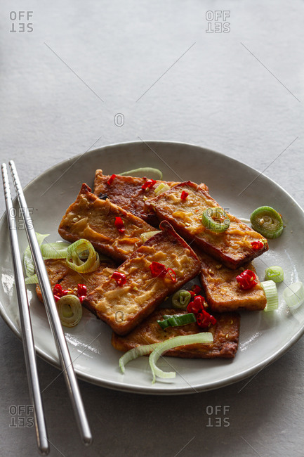 Top view of tofu slices with fragrant pieces of garlic marinating in soy sauce in white deep plate while standing on kitchen towel on gray table with steel chopsticks