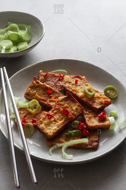 Top view of tofu slices with fragrant pieces of garlic marinating in soy sauce in white deep plate while standing on kitchen towel on gray table with steel chopsticks
