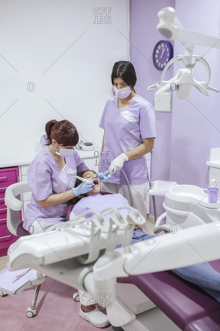 Female dentist in uniform and mask curing patient teeth with female assistant in modern dental clinic