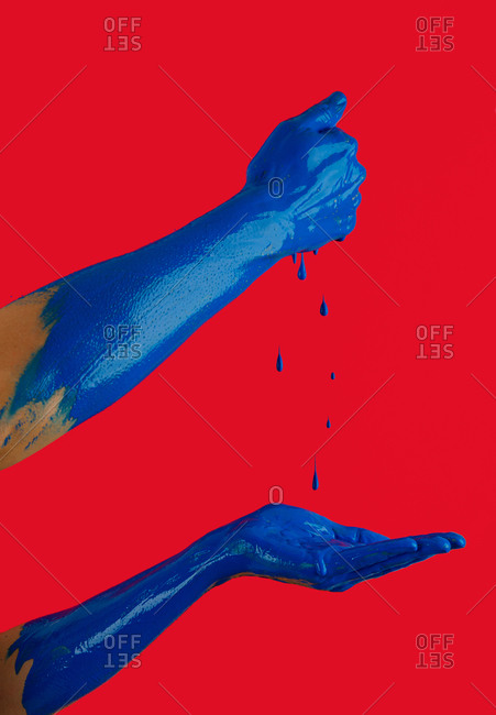 Unrecognizable person with painted hands squeezing blue droplets of gouache on red background in studio