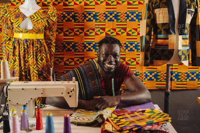 Happy black male tailor creating national costume of colored fabric with African patterns on sewing machine while sitting on background of colored African material and mannequin in sewing workshop