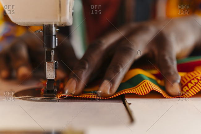 Crop male tailor creating national costume of colored fabric with African patterns on sewing machine while sitting on background of colored African material and mannequin in sewing workshop