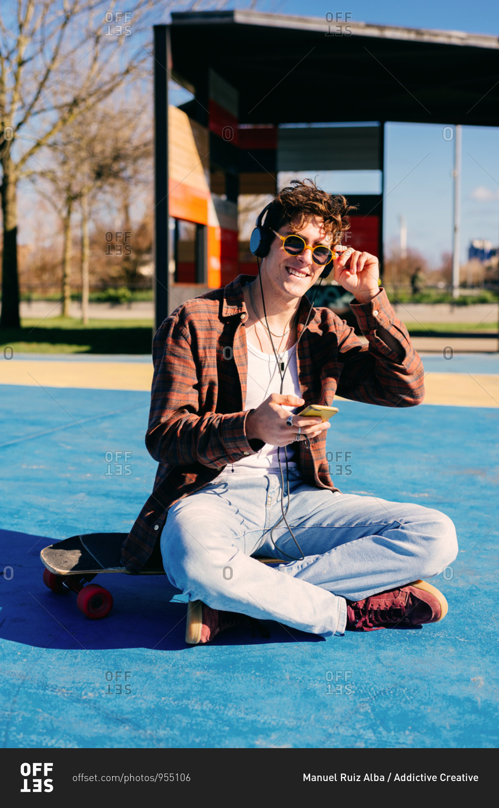Happy hipster in casual clothes smiling for camera and browsing smartphone while sitting on skateboard and listening to music on sports ground