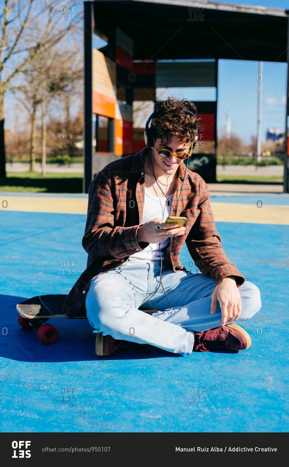 Happy hipster in casual clothes smiling and browsing smartphone while sitting on skateboard and listening to music on sports ground