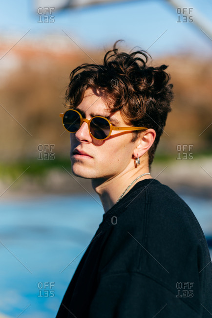 Handsome young man in dark t-shirt, sunglasses and earrings looking at camera over the shoulder while standing on blurred background of city street