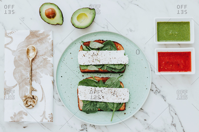 From above plate with cheese and spinach sandwiches place on marble tabletop near napkin with spoon and bowls with avocado and tomato sauces