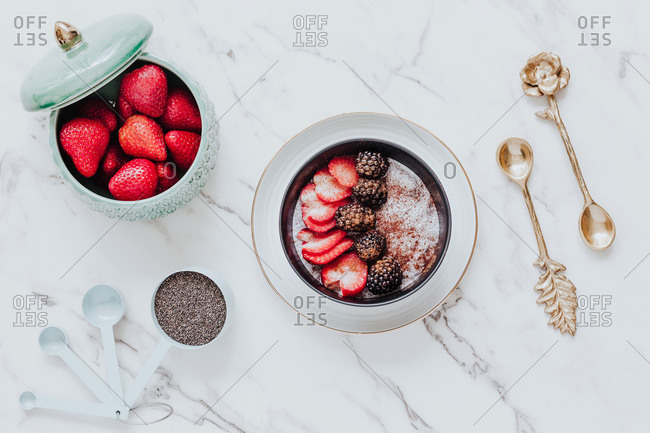 From above slices of ripe strawberries and blackberries in cinnamon powder in yogurt bowl in composition with bowl with fresh whole berries and golden teaspoons on white table