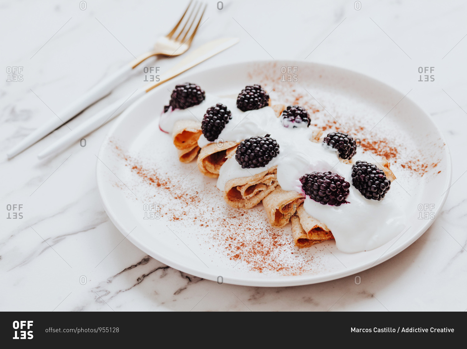 Delicious crepes with yoghurt and blackberries served on plate with cinnamon near silverware on marble table
