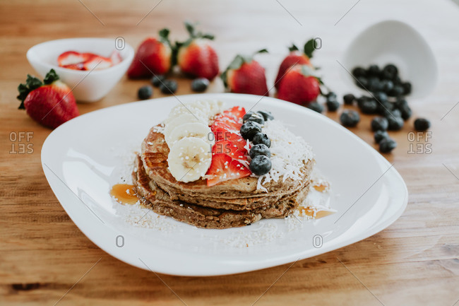 Stack of tasty pancakes served on plate with pieces of banana and strawberry and fresh blueberries with coconut flakes during breakfast