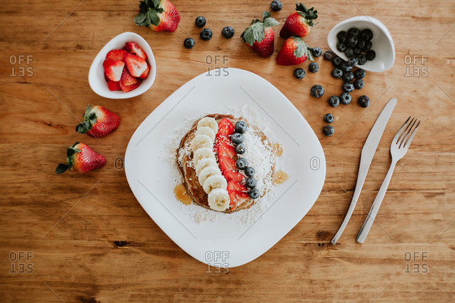Stack of tasty pancakes served on plate with pieces of banana and strawberry and fresh blueberries with coconut flakes during breakfast
