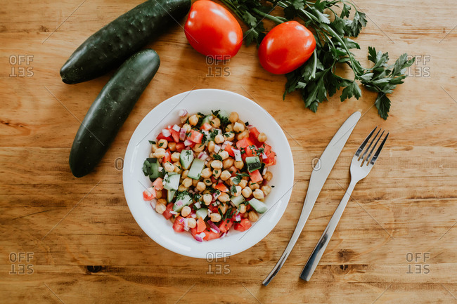 From above bowl of healthy cucumber and tomato salad with chickpeas and parsley placed on lumber table near fork and knife