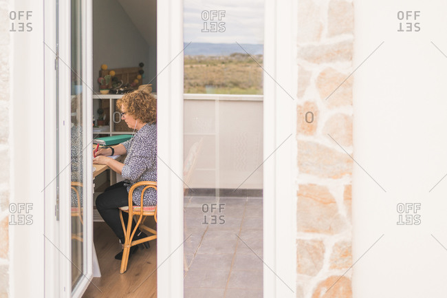 Through window side view of serious adult woman in casual clothes and eyeglasses taking notes with pen in notebook while working in cozy chair at table at home