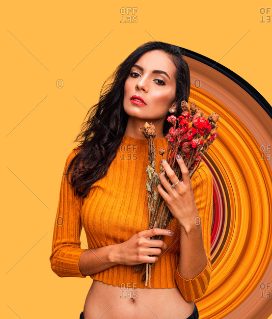 Confident young female model in trendy orange crop top holding bouquet of fresh flowers and looking at camera while standing against bright orange wall with geometric ornament in studio