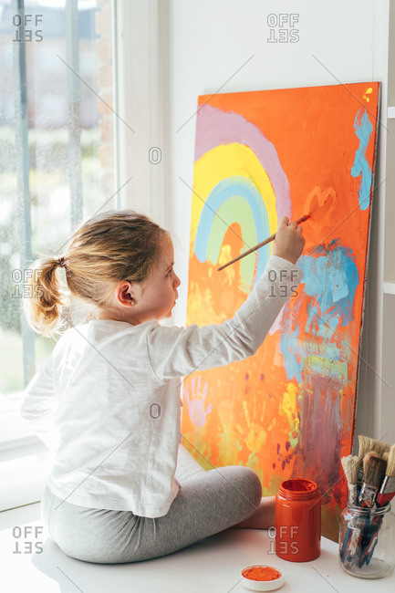 Creative blond girl in casual clothes sitting on window sill against window and painting with paintbrush large multi colored rainbow on orange canvas