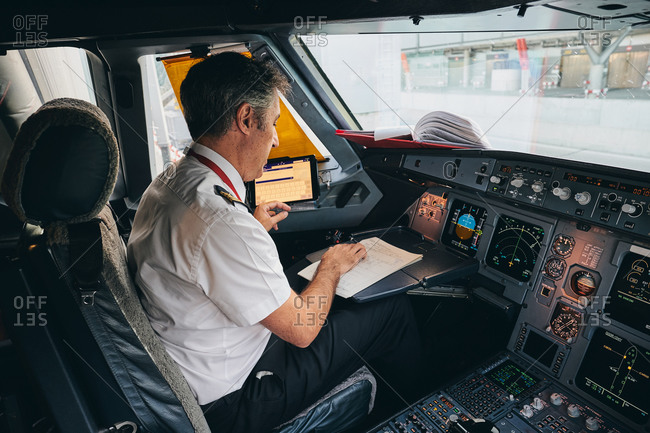 Side view of male captain sitting in cockpit of contemporary aircraft and reading flight document while preparing for departure