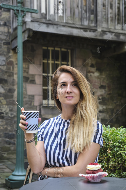 Dreamy female in casual wear taking hot drink from disposable cup while sitting at round table with cupcake garnished with buttercream and raspberries in street cafeteria in sunlight