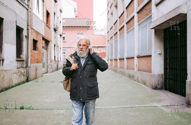Senior male with long gray hair looking away using mobile phone while walking on the street surrounded by shabby buildings