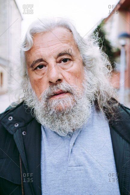 Serious senior male pensioner in casual wear smiling at camera while standing on blurred street in city at daytime