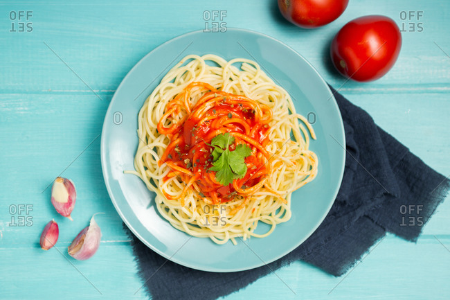 Top view of blue ceramic plate with pasta and tomato sauce decorated with parsley and basil served between cloves of garlic and couple of tomato on light blue background