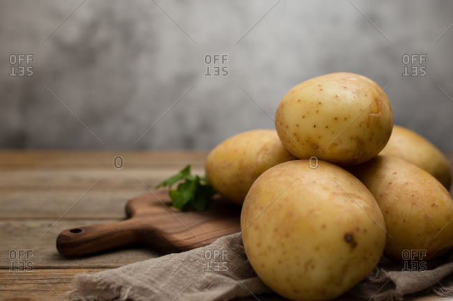 Raw potatoes placed on wooden cutting board near linen cloth and green parsley on wooden table against gray background