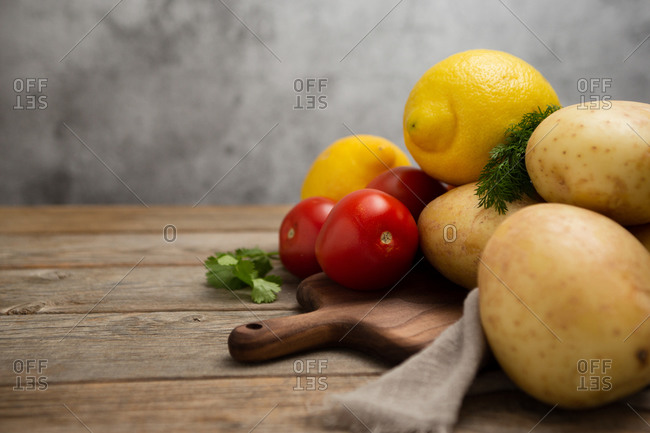 Composition of different raw vegetables placed on wooden cutting board near linen cloth and various greens on wooden table against gray background