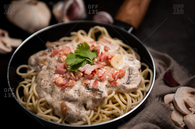 Composition of tasty spaghetti with ham slices and mushrooms in creamy sauce cooked in pan and placed on wooden cutting board at dark table with garlic and gray linen fabric aside